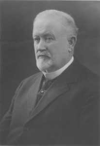 Studio photograph of a man with white hair and wearing a black suit.