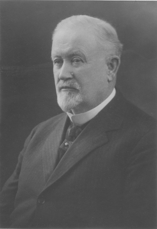 Studio photograph of a man with white hair and wearing a black suit.