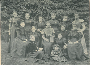 14 women standing outside in front of trees.