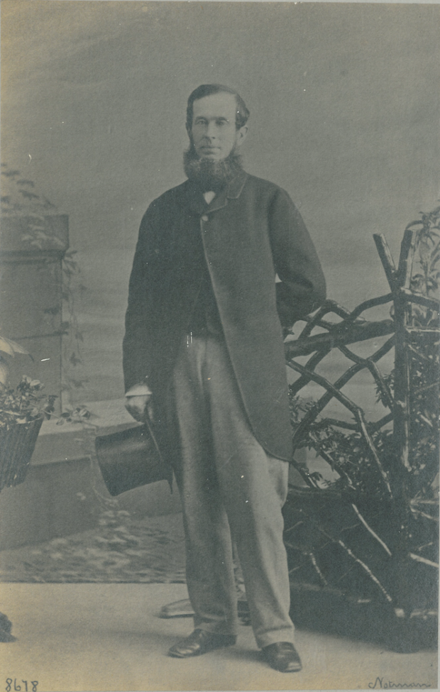 Studio photograph of a man standing while holding a top hat by his waist.