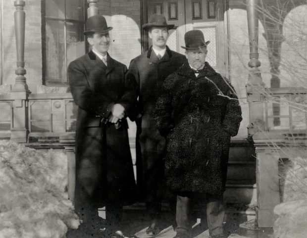 Three men standing on front steps wearing overcoats during winter.