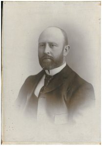 Studio photograph of a middle aged man wearing a black suit with a white shirt.