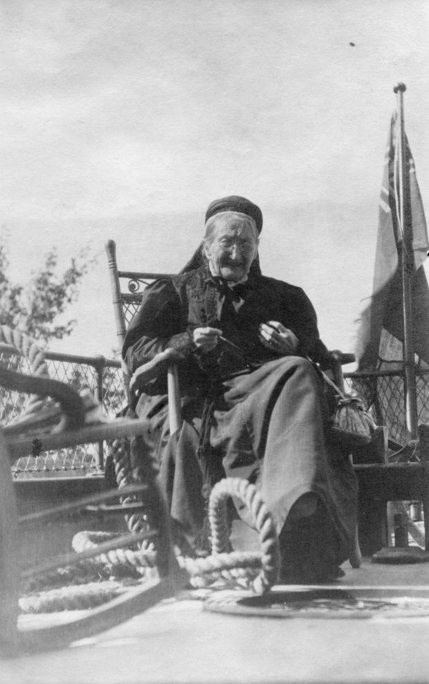 Old woman knitting in a chair on the top deck of a house boat wearing a black dress.