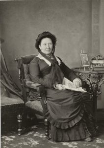 Studio photograph of a woman wearing a black dress sitting in a chair.