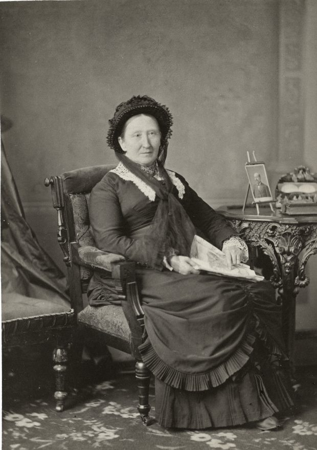 Studio photograph of a woman wearing a black dress sitting in a chair.