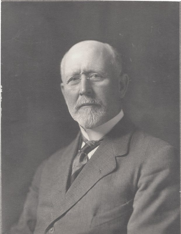 Studio photograph of man with white hair and glasses wearing a black suit.