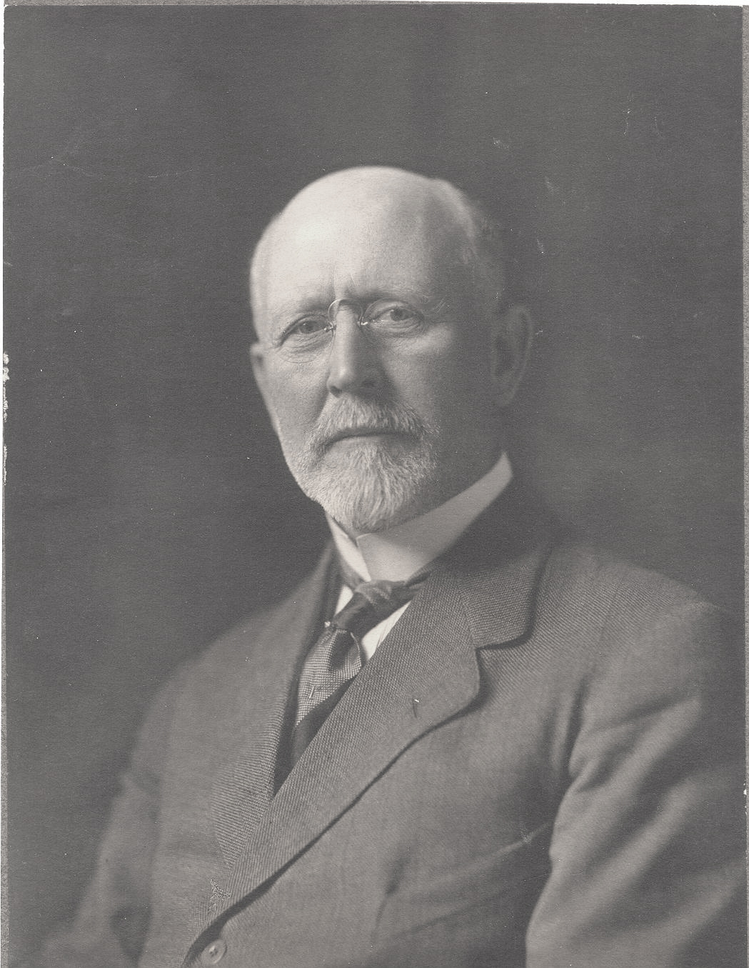 Studio photograph of man with white hair and glasses wearing a black suit.