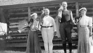 Two women and two men holding golf clubs in front of a log cabin.
