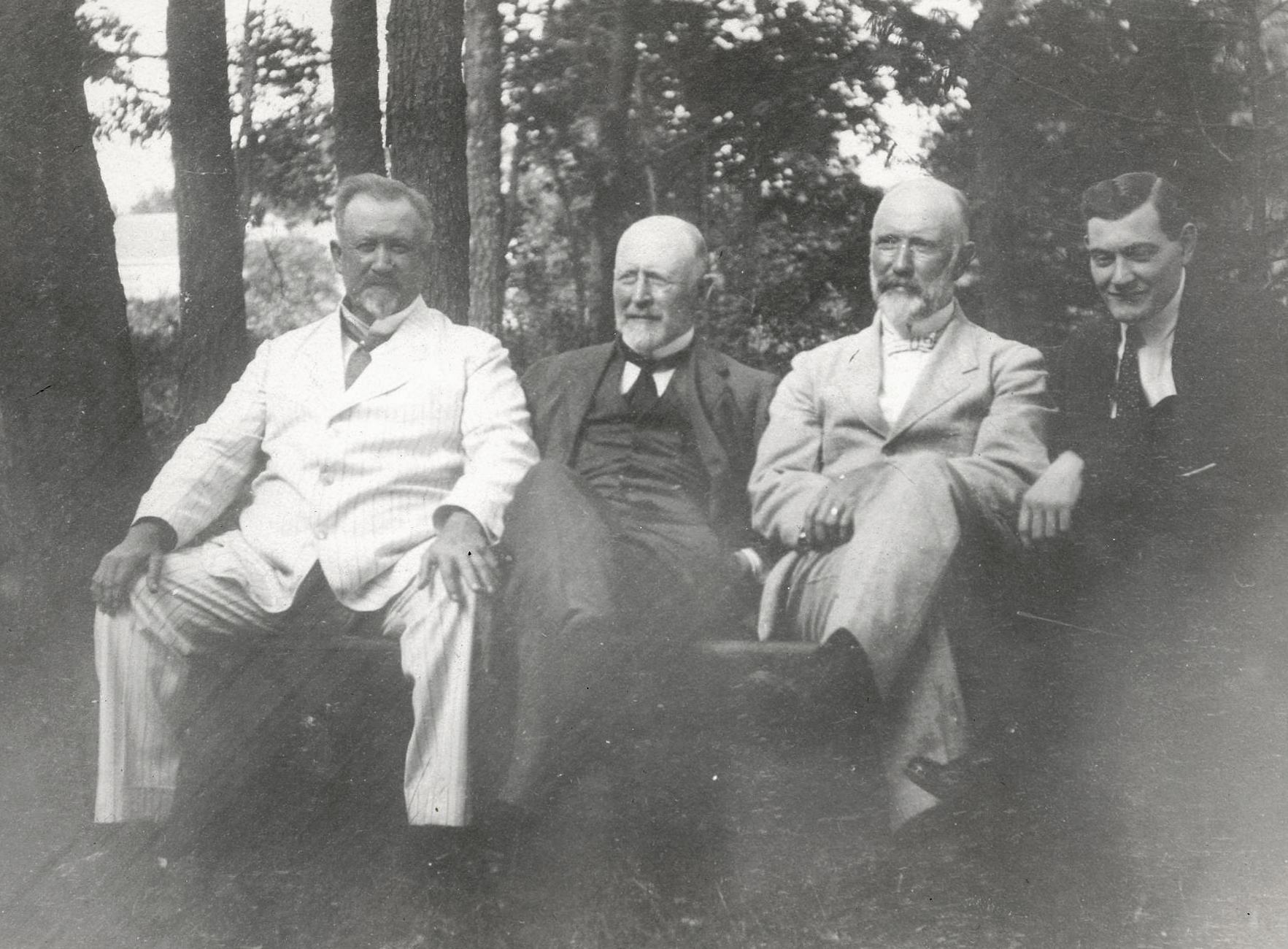 4 men sitting on a bench in front of trees.