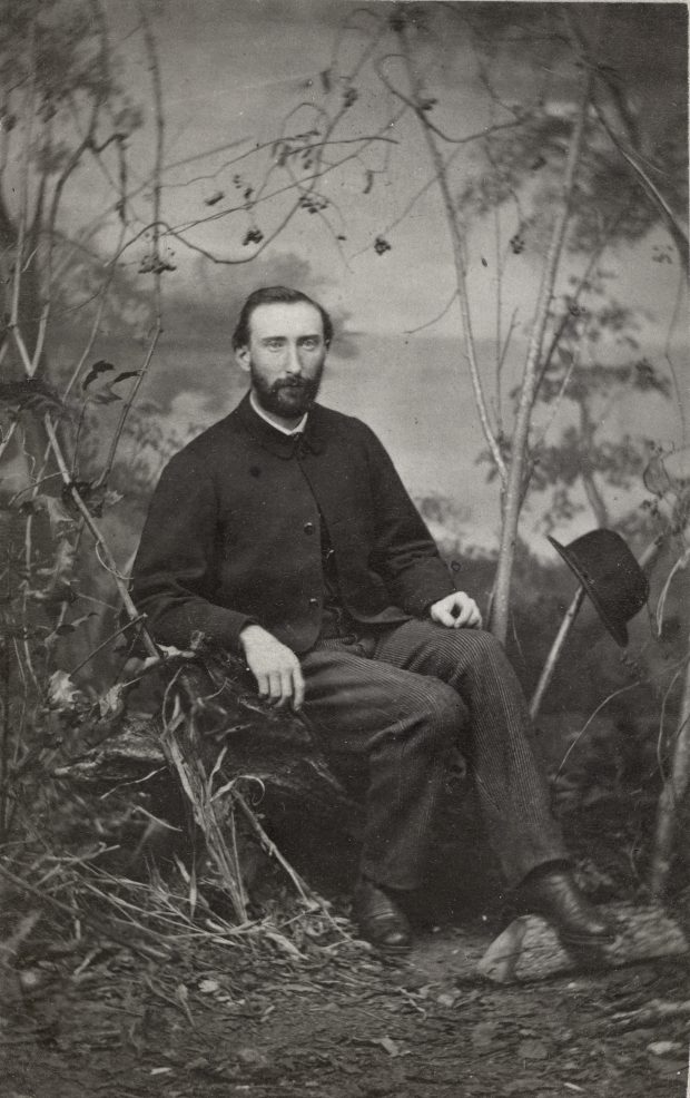 Studio photograph of middle aged man seated by shrubbery.