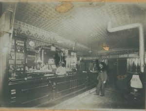 A bartender in a white coat behind a bar serving a customer who is wearing a black suit and hat in a hotel bar.