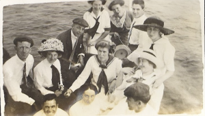 Group of young men and women dressed in white dresses and black suits in a boat on water.