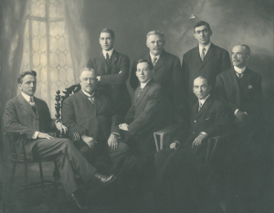 Studio photograph of eight men wearing suits.