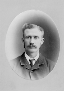 Studio photograph of a young man with combed hair and a moustache wearing a black suit.