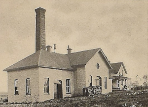 Brick building with a tall smoke stack at the rear of the building with cut wood at the front.