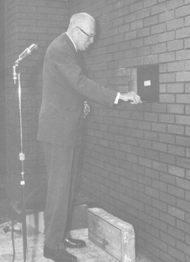 Man with glasses standing behind a microphone about to lay a stone in a brick building