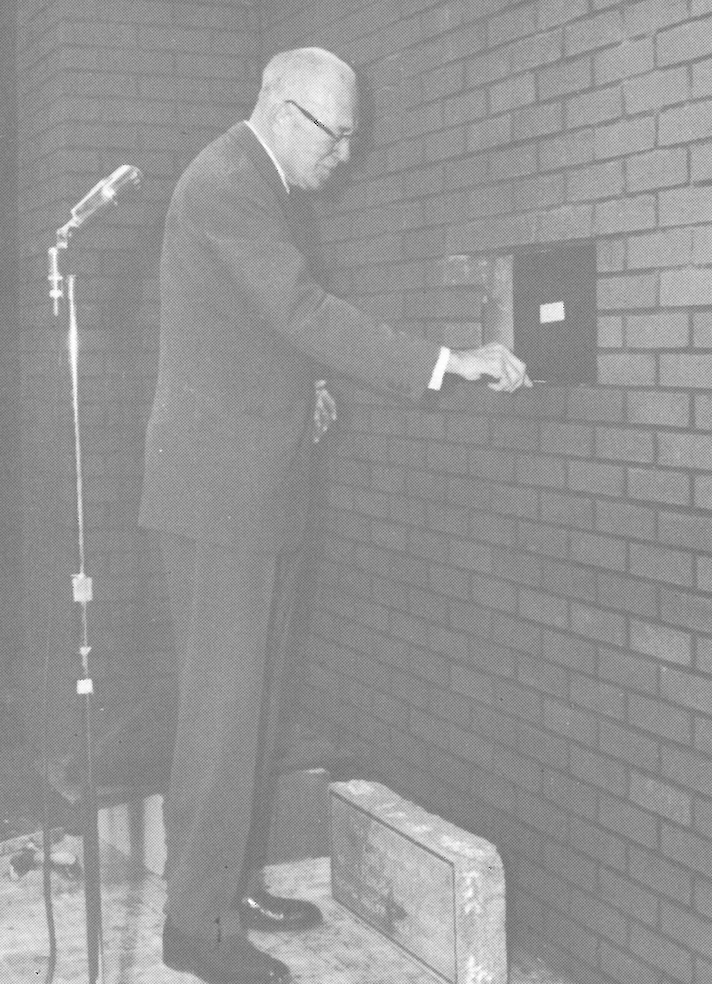 Man with glasses standing behind a microphone about to lay a stone in a brick building