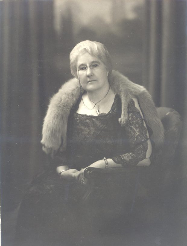 Woman wearing glasses with grey hair in a black lace dress and a fox pelt around her neck sitting in a chair.
