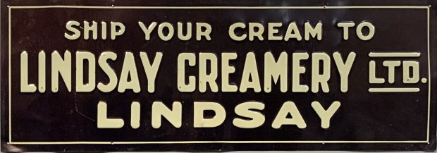 Metal sign painted black with Ship Your Cream To Lindsay Creamery Ltd. LINDSAY painted in bold yellow print.