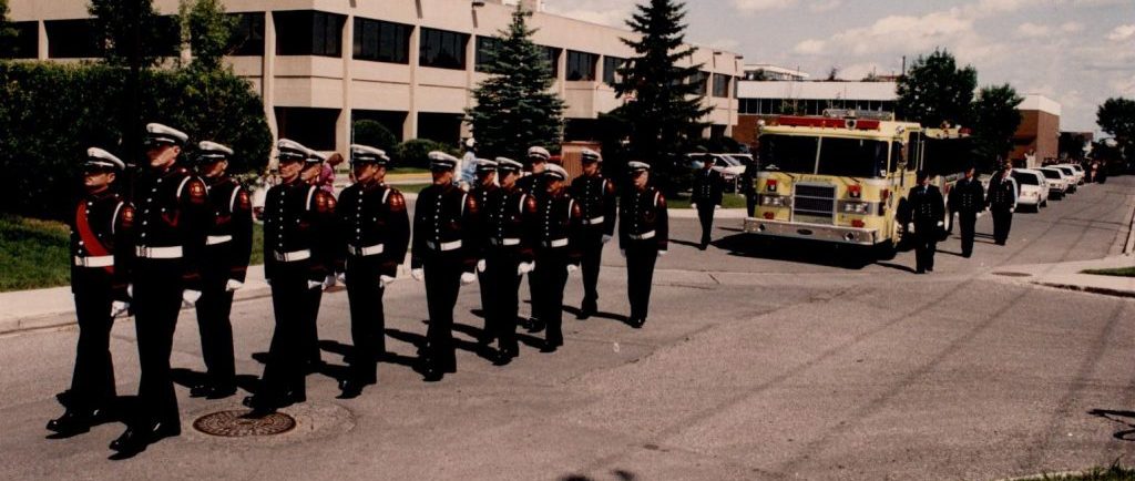 Several members of the Guard of Honor walk in uniform in front of a yellow fire truck at the head of Morley James' funeral procession.