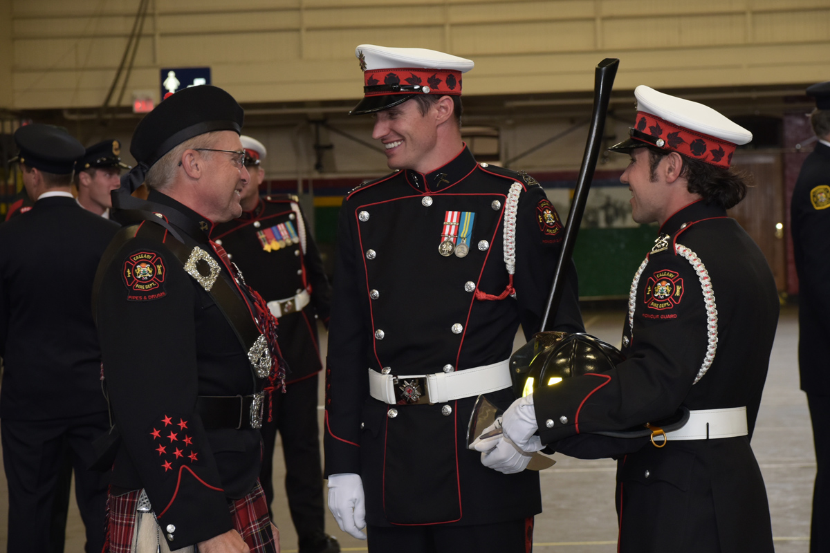 Two Honour Guard Members and a piper from Pipes and Drums, all in full uniform, laugh as they talk together.