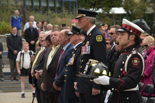 Guardsman John Judge in full uniform and white gloves holds black ceremonial helmet with yellow patches at Tribute Plaza with chief and crowd in background.