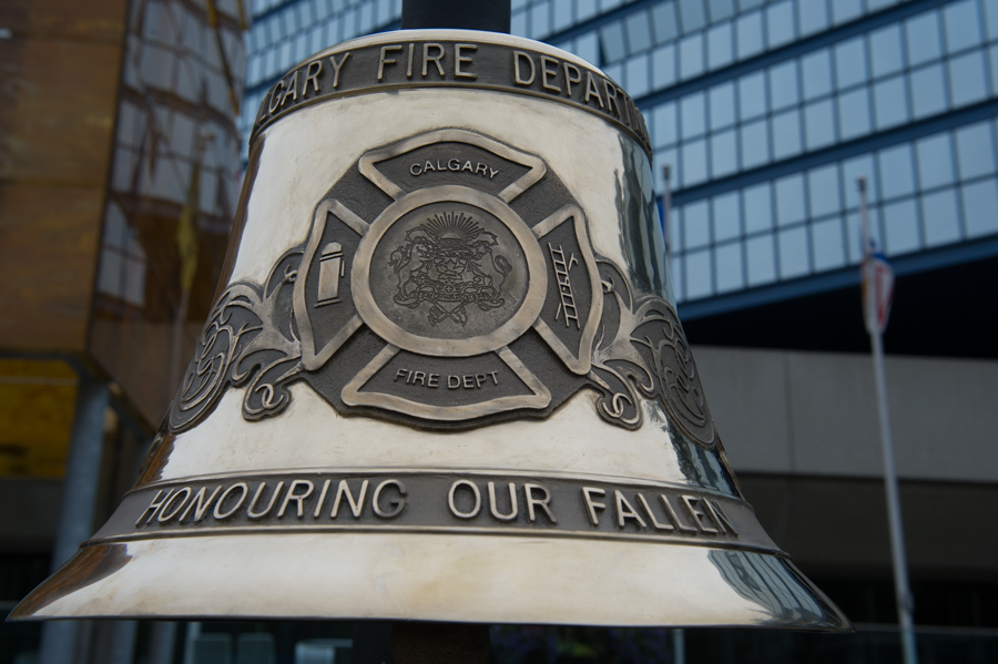 Close up on silver bell inscribed Calgary Fire Department and Honouring Our Fallen with CFD crest. City hall can be seen in the background.