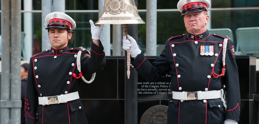 A large, hanging bell is held and rung by two Honour Guards in full dress uniform and white gloves, in front of the memorial at Tribute Plaza, City Hall