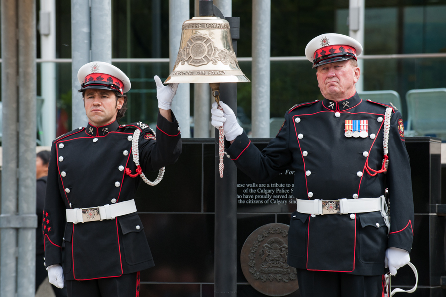 A large, hanging bell is held and rung by two Honour Guards in full dress uniform and white gloves, in front of the memorial at Tribute Plaza, City Hall
