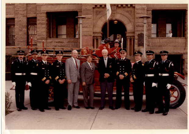 Eight guard members stand with former mayors, in suits, in front of antique fire truck and sandstone city hall.
