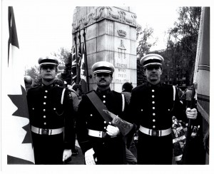 Three Honour Guard bearing flags and a ceremonial axe stand in front of the WWI Memorial at Central Memorial Park. Photo is black and white.