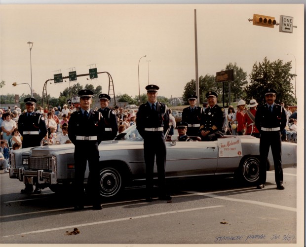 Six Honour Guard members are standing around a convertible with sitting Fire Chief. In the background are parade goers lining the street.