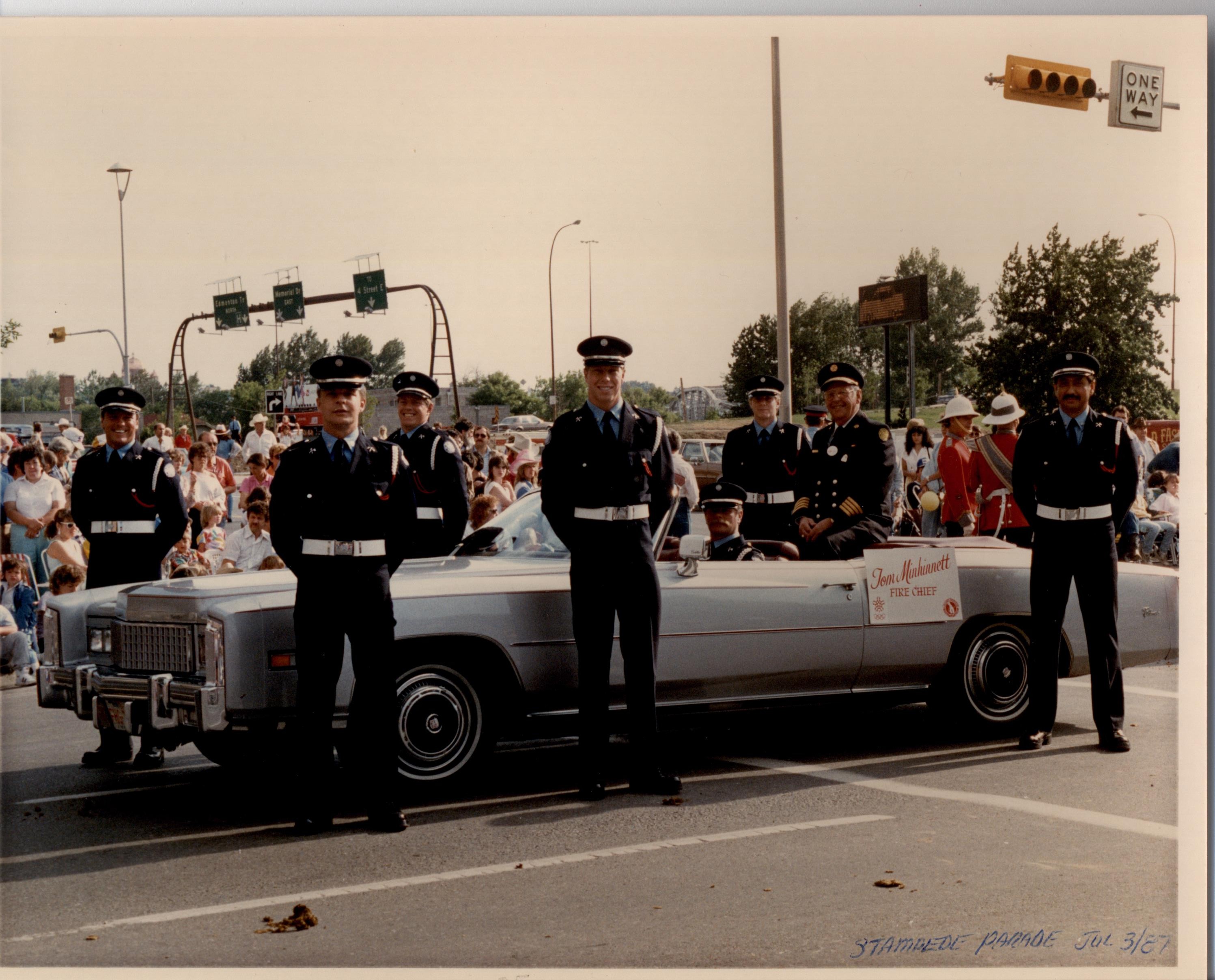 Six Honour Guard members are standing around a convertible with sitting Fire Chief. In the background are parade goers lining the street.