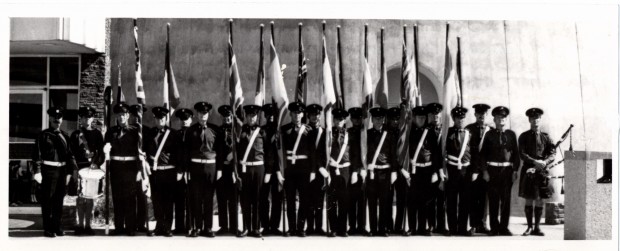 Black and white photo showing more than 20 men standing in a line at attention in full uniform at the base of the tower. Most carry flags. Men at either end are carrying pipes and drums