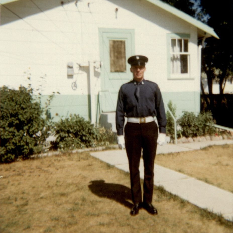 Dennis McIvor, standing on lawn in front of white and blue house, dressed for Honour Guard duty, in original Number 1 dress with tie tucked in.