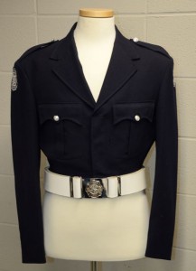 Original, Number 1 dress Honour Guard uniform. Black battle style jacket with CFD patch on both shoulders, two breast pockets, box pleated, with scalloped flaps and two epaulettes secured by silver buttons. White leather belt and silver plated buckle over the jacket on a white headless mannequin.