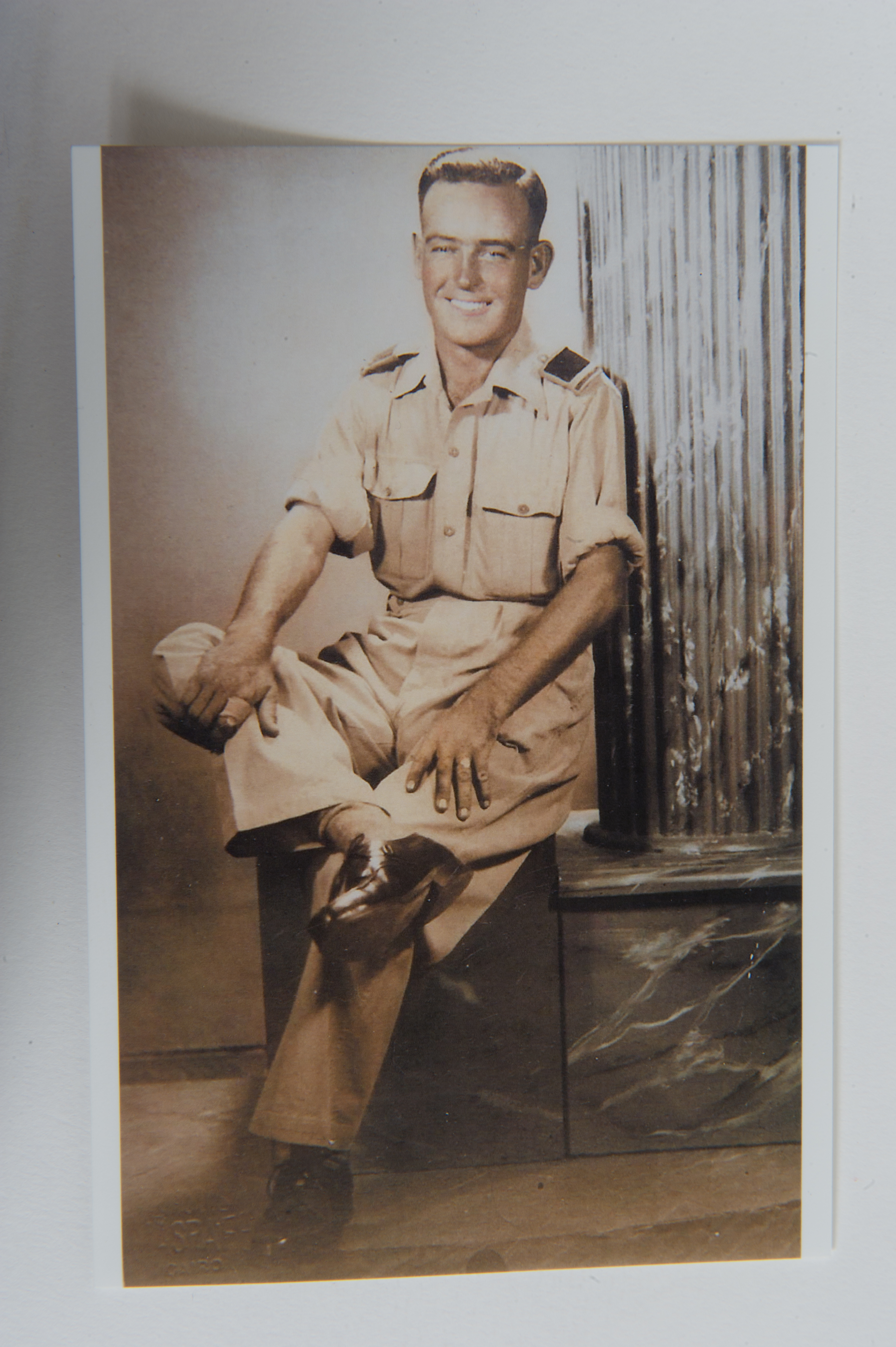 A sepia toned photo shows a man in military uniform, sitting causally with right leg crossed over left knee, shirt sleeves rolled up, and both hands on knees.