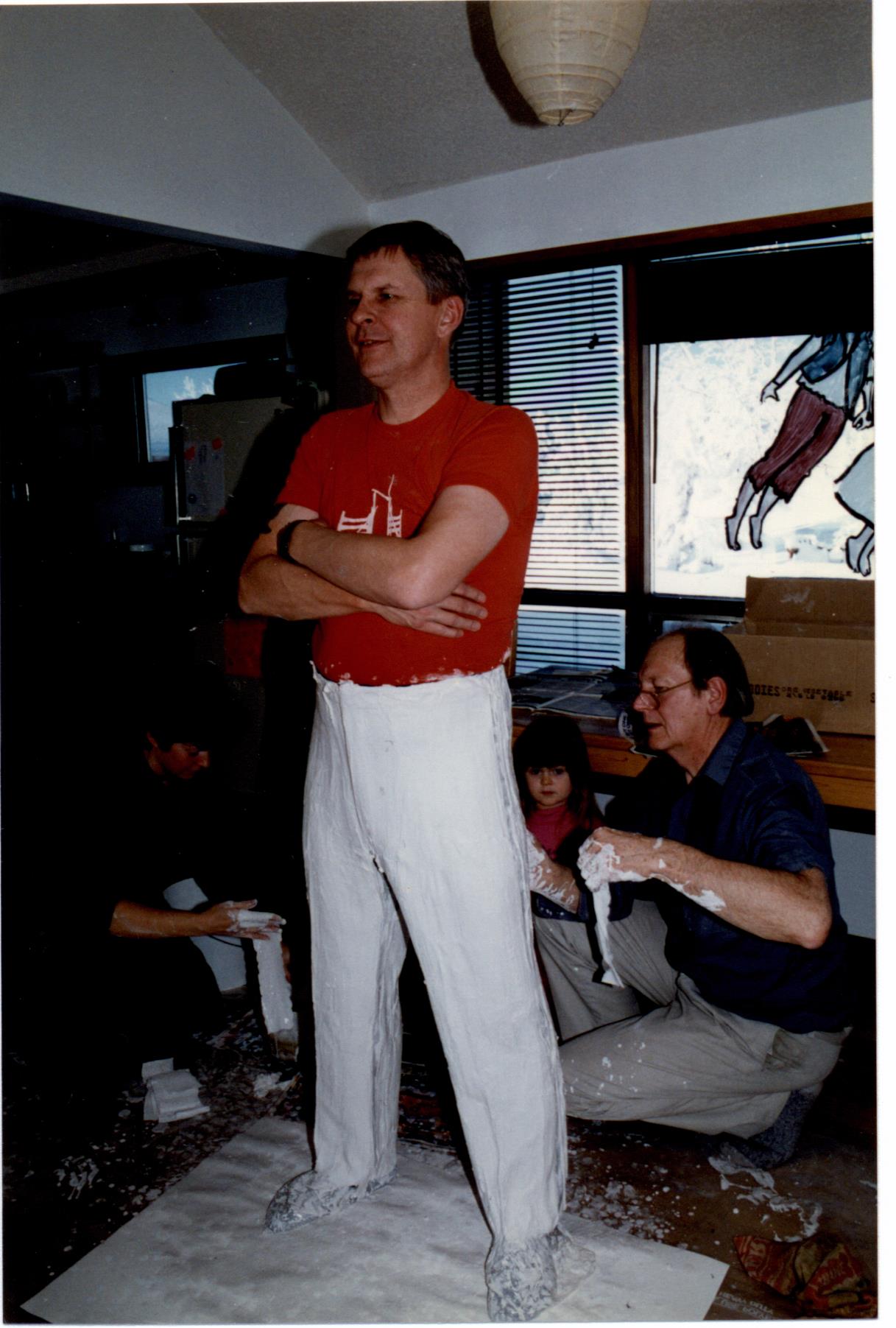 Rick Choppe stands still with arms crossed as plaster is applied to his legs by an artist, crouching.