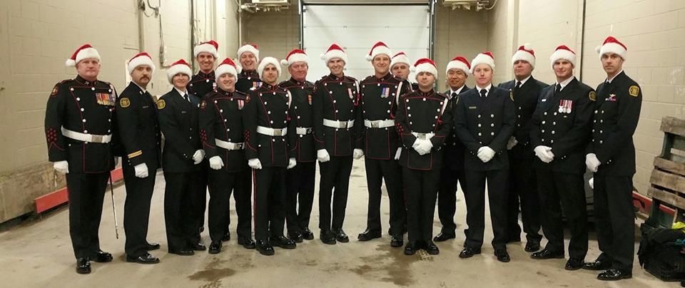 Seventeen firefighters in dress uniform all stand in a line, smiling backstage, wearing red Santa hats