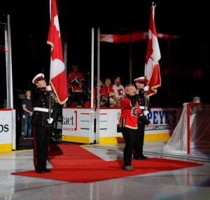 Two Honour Guard members stand at attention in full dress each carrying the Canadian Flag while blonde singer sings the National Anthem in a Calgary Flames jersey.