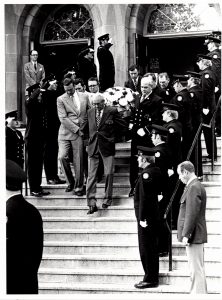 A funeral cask held by pallbearers leaves a church; the path is lined by uniformed and saluting firefighters.
