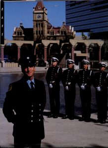 A firefighter stands in the left foreground, with four Honour Guard members standing at attention mid ground. In the background is Olympic Plaza and historic sandstone city hall.