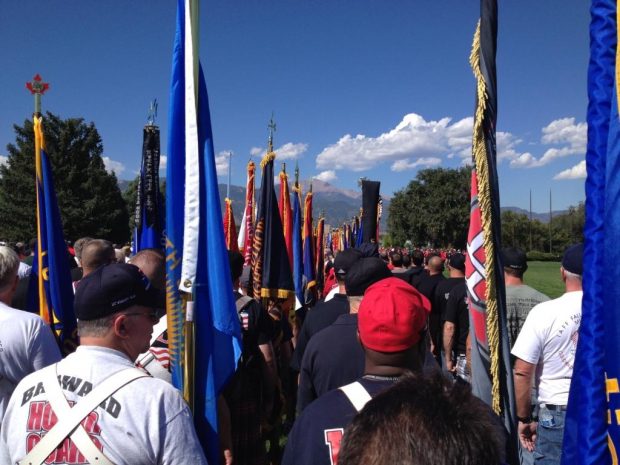 Honour Guards in casual dress prepare to march in memorial procession, with backs to camera, holding colourful flags. In the background are mountains.