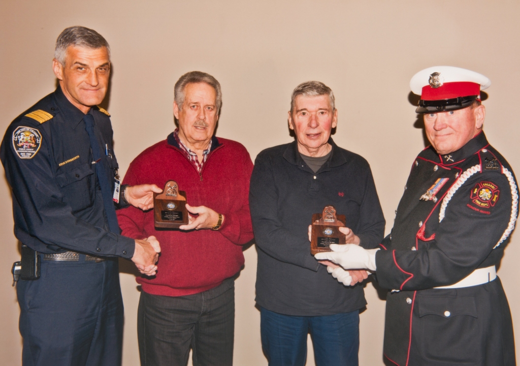 Founders Brian Freney and Dennis Mcivor, in casual outfits, are presented with their commemorative plaques by two uniformed firefighters.