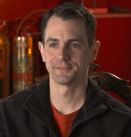 Head and shoulder colour photo of Derek Rogers wearing a red tee-shirt and a black golf jacket.