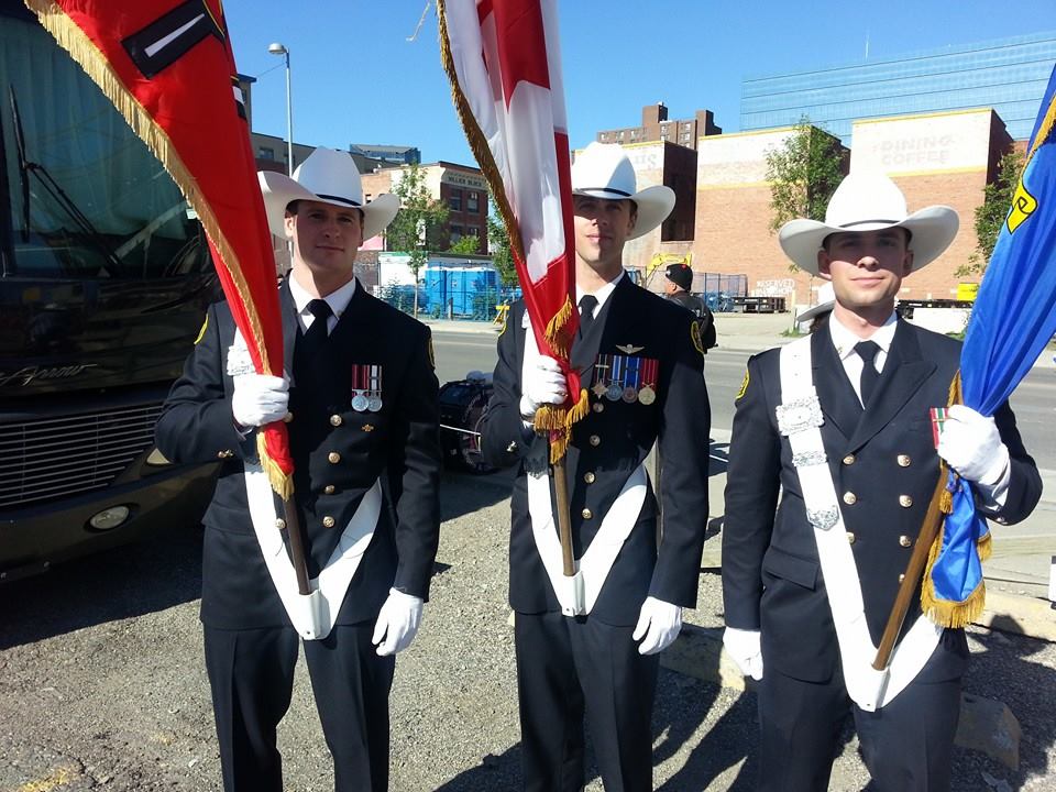 Three smiling Honour Guards in uniform with medals, wearing white cowboy hats and holding flags, prepare to march in the parade. It's a sunny day with city buildings in the background.