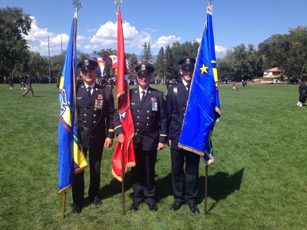 Three Honour Guard members stand in full uniform in a green, grassy field, each holding a different flag.