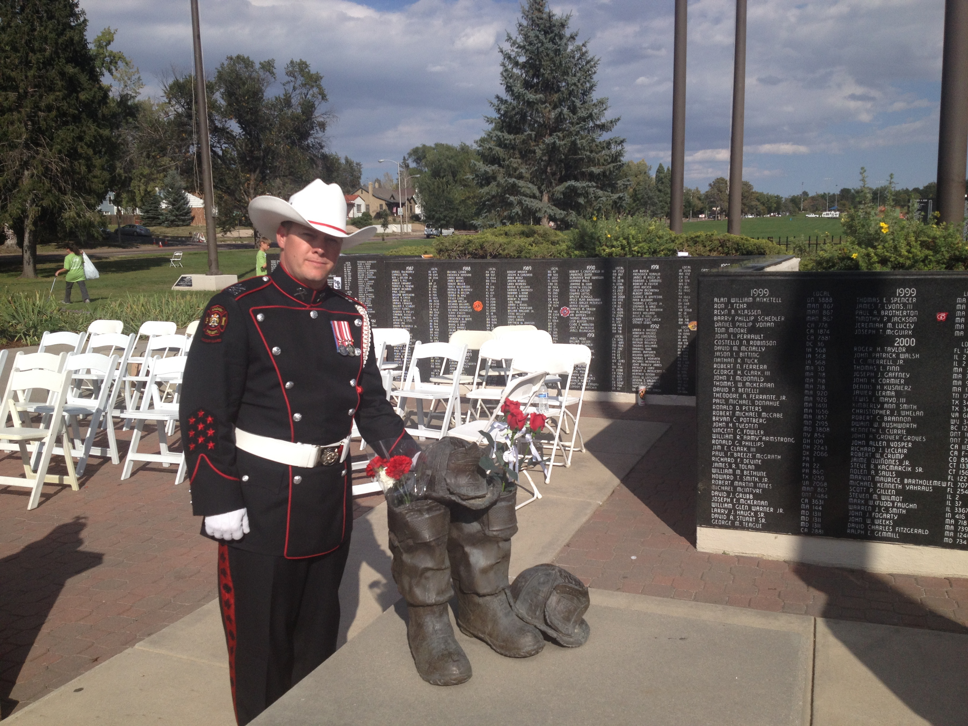 A CFD Honour Guard in unifrom with white belt, gloves and cowboy hat stands with a sculpture of boots, in front of a memorial wall inscribed with names.