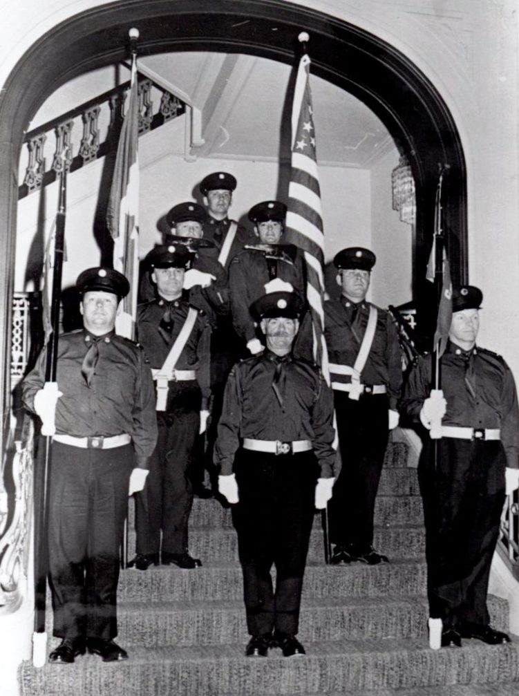 Black and white photo of eight members of the Honour Guard stand at attention on a set of stairs underneath an arch at their first engagement in 1971.
