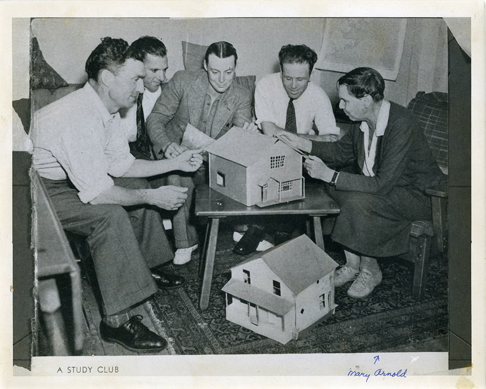 Black and white photograph of a group of men and women in a study group with model houses. Hand written at the bottom 